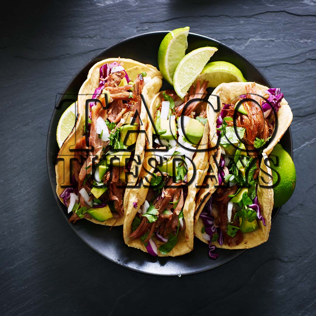 Taco Tuesdays - three tacos on a black plate with avocado and lime wedges